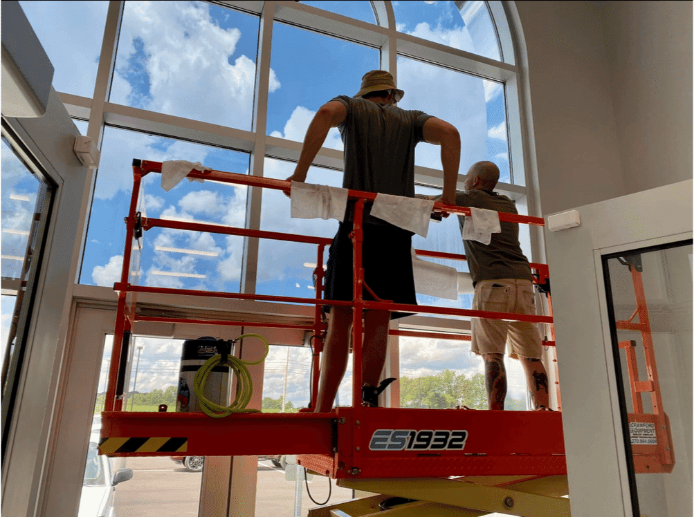 Two workers on a lift cleaning large windows with blue sky and clouds visible outside.