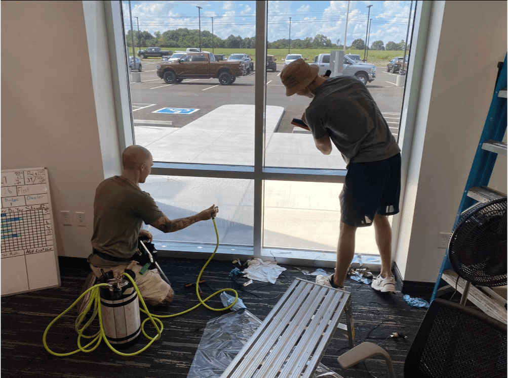 Two men cleaning a large window with tools and equipment, bright outdoor setting visible.