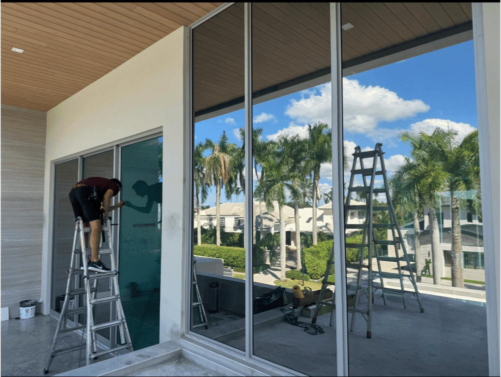 Worker cleaning large glass windows with ladders in modern home surrounded by palm trees.