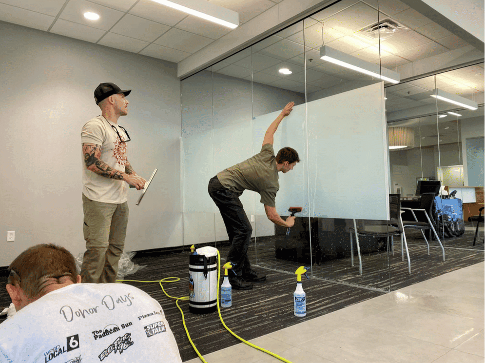 Two men cleaning glass office partitions with a spray bottle and squeegee in a modern workspace.