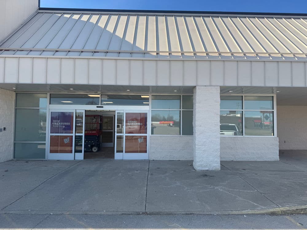 Entrance of a retail building with glass doors and a light gray facade, bright day outside.